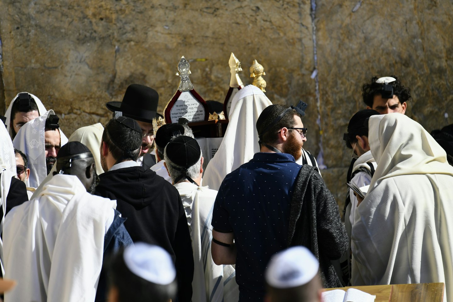 Men in religious attire hold Torah scrolls near a stone wall.